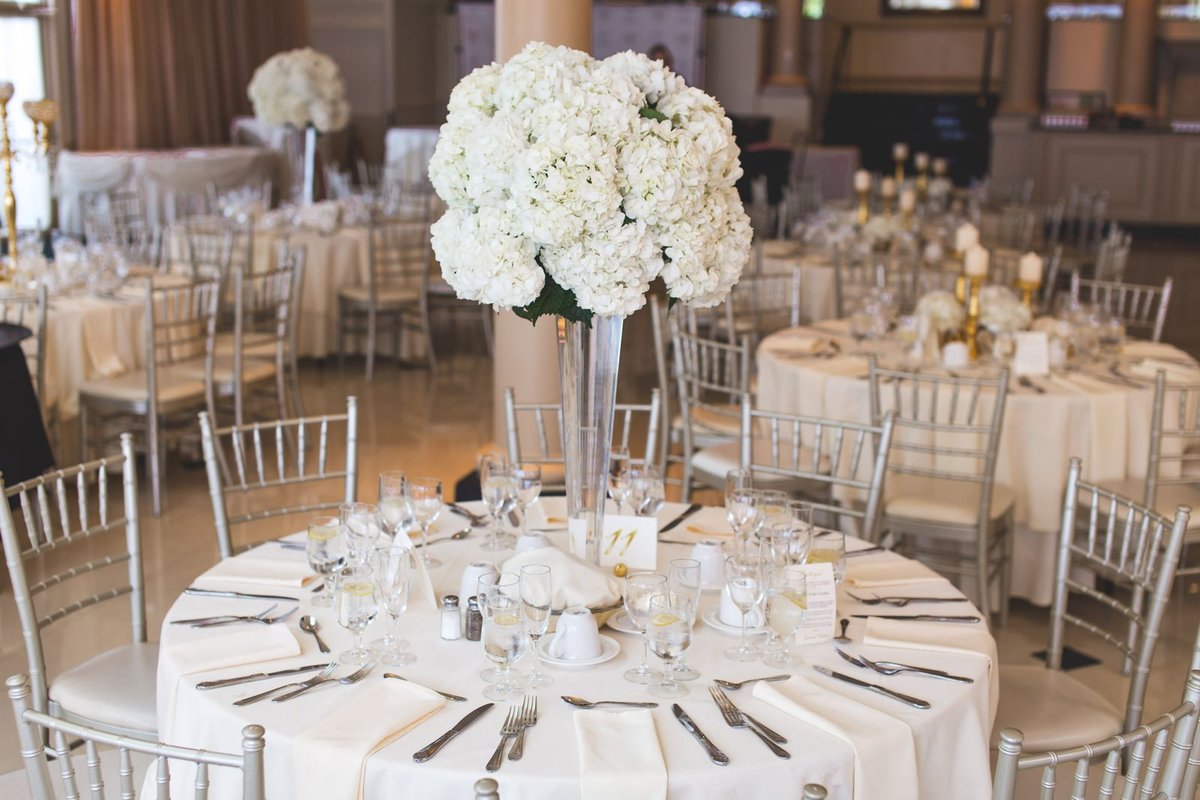Round banquet table with white floral centerpiece, silver Chiavari chairs, and place settings in a bright event hall