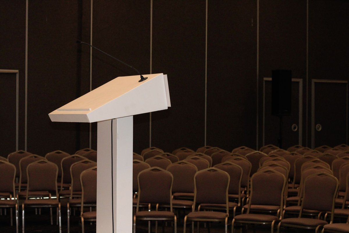 White lectern with gooseneck microphone facing empty rows of dark chairs in a dim conference hall